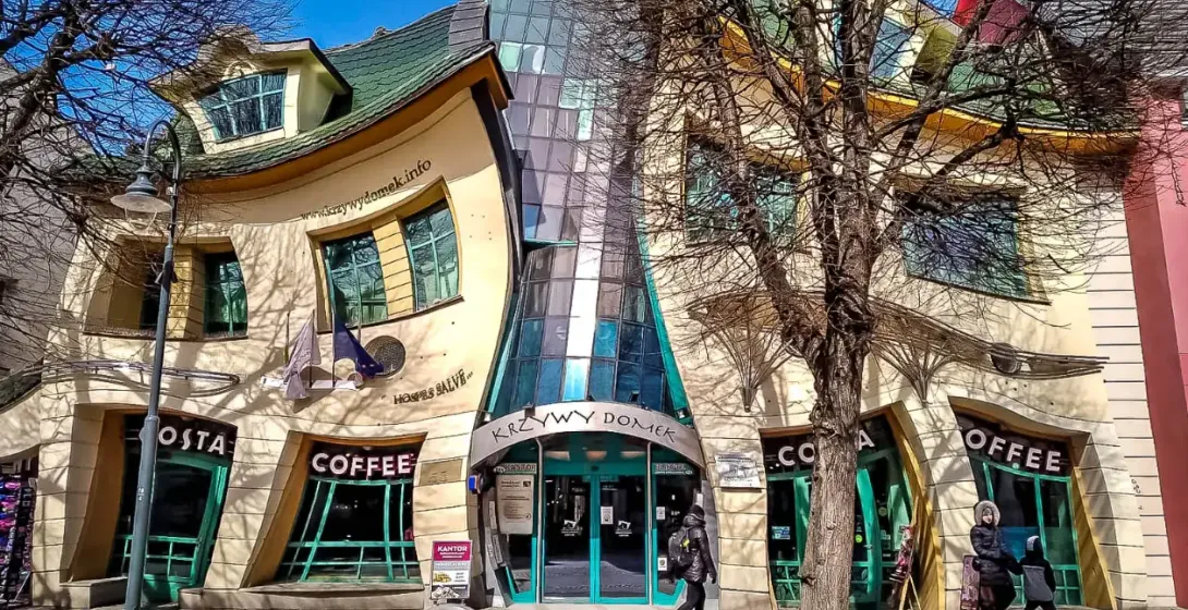 The Crooked House (Krzywy Domek) in Sopot, with a wavy facade and green roof, shops on the ground floor, and bare trees.