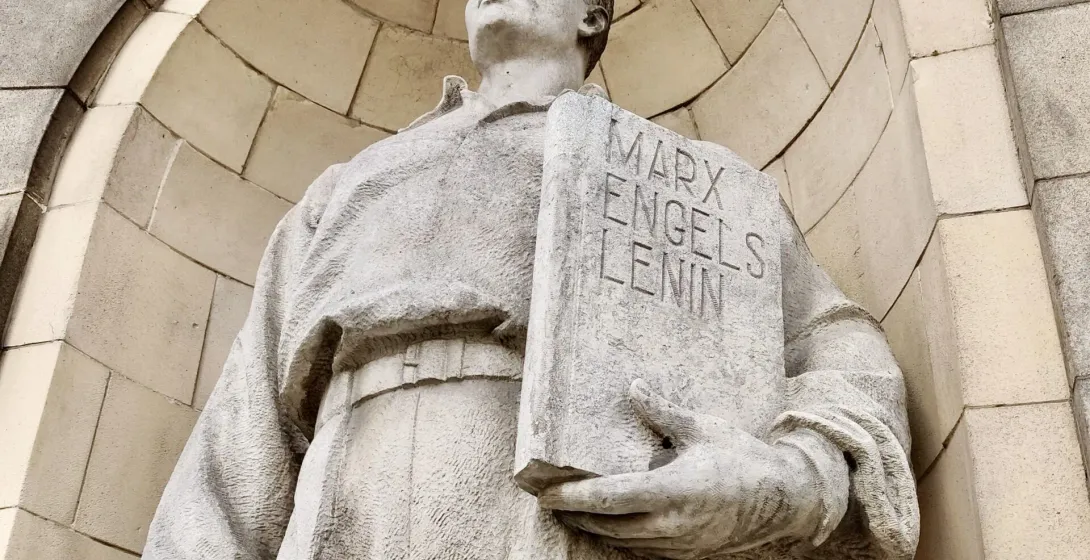 Close-up of a weathered stone statue holding a book inscribed with 'Marx Engels Lenin'.