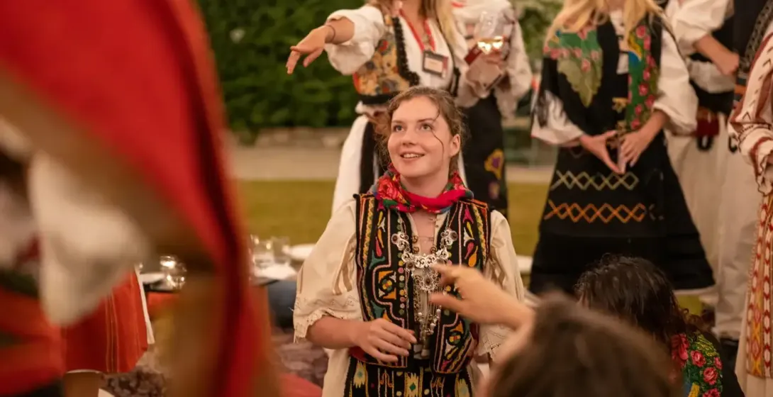 Young woman in embroidered traditional Albanian clothing and a red scarf smiling, with other people in traditional attire ...
