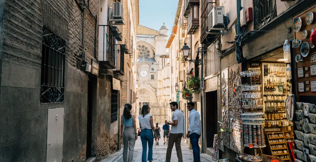 Four young people walk down a narrow alley in Toledo, with the cathedral in the background and a souvenir shop on the right.