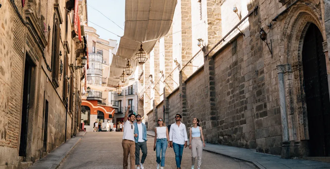 A group of five young people walks down a narrow stone street in Toledo, shaded by traditional awnings.