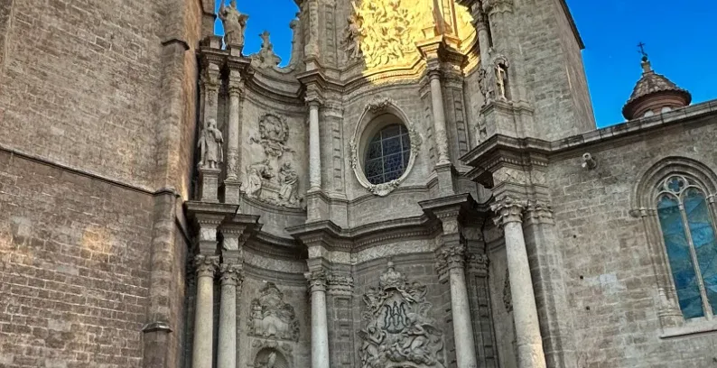 Detailed baroque facade of Valencia Cathedral with a prominent tower and entrance, bathed in sunlight against a deep blue ...