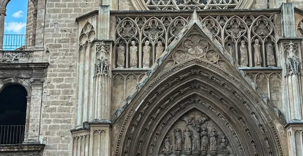 Gothic stone facade of Valencia Cathedral with a large rose window, ornate arched portal, and sculpted figures under blue ...