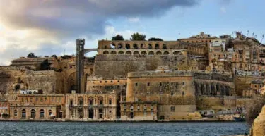 View of Valletta's historic fortifications and stone buildings along the Grand Harbour, with a dramatic cloudy sky above.