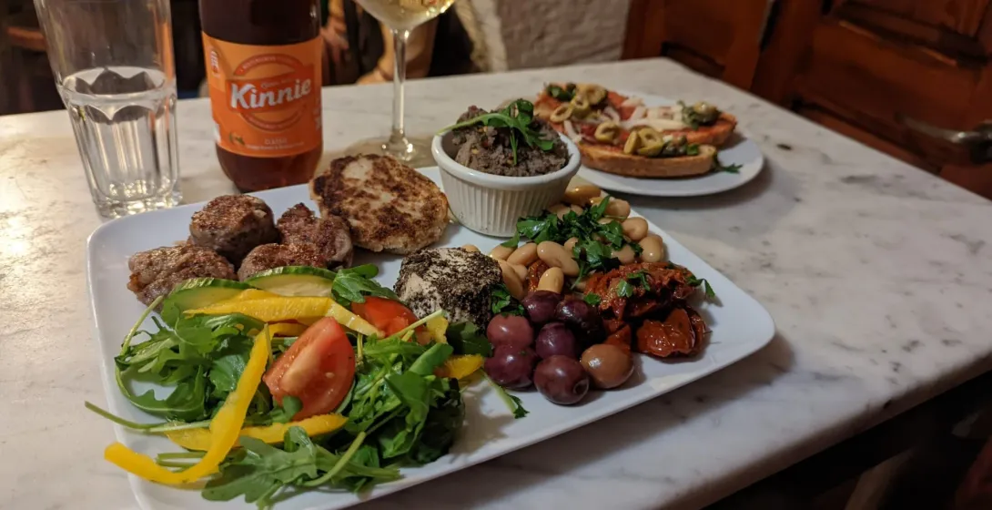 A platter of Maltese food including meatballs, salad, beans, olives, and cheese, with a Kinnie bottle and wine glass on a ...