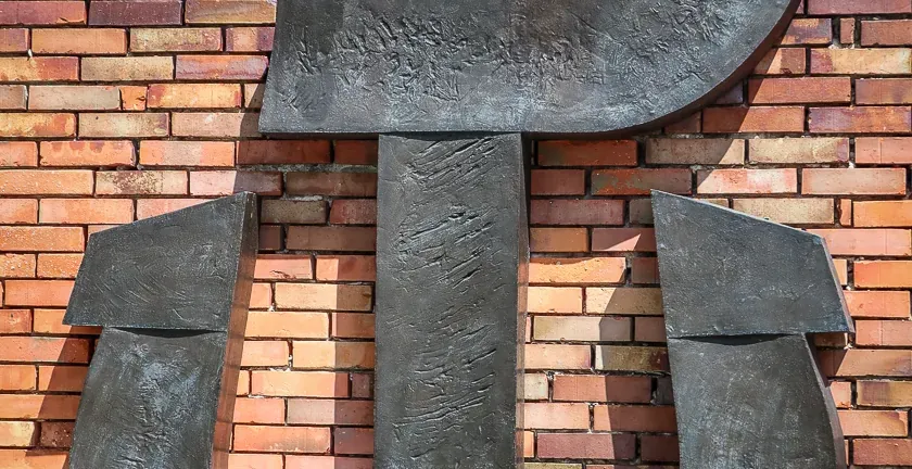 Large, dark metal sculpture of the Fighting Poland (Kotwica) symbol on a multi-colored brick wall with green leaves above ...