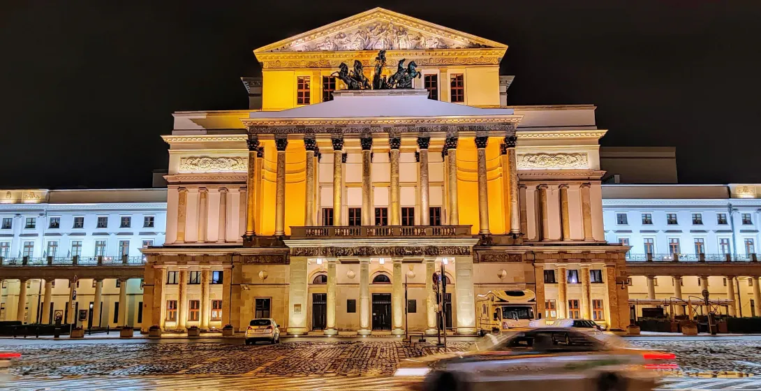 Grand Theatre in Warsaw at night, with a classical facade, columns, and sculptures, illuminated by warm lights. Snow on th...