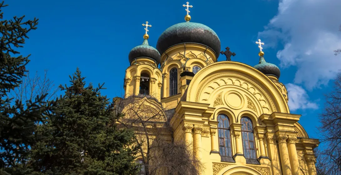 Yellow Orthodox church with dark domes and golden crosses against a blue sky, surrounded by green trees.