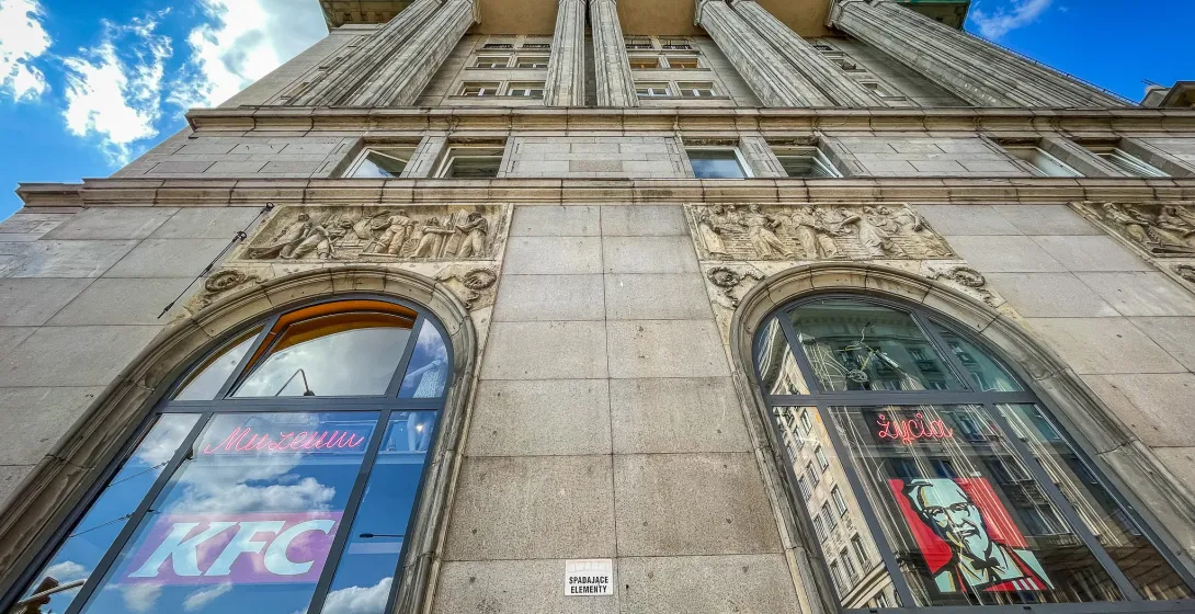Low-angle view of a grey stone building with classical columns and relief carvings. Large arched windows show reflections,...