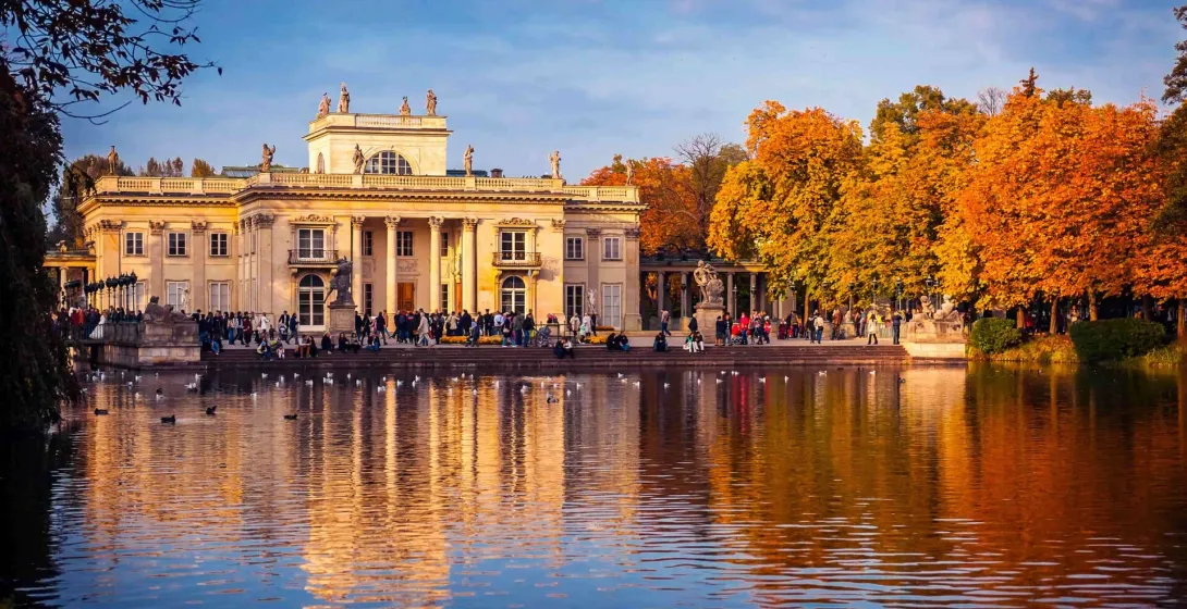 Palace on the Isle at Łazienki Park in Warsaw, reflected in a pond with ducks, surrounded by autumn trees.