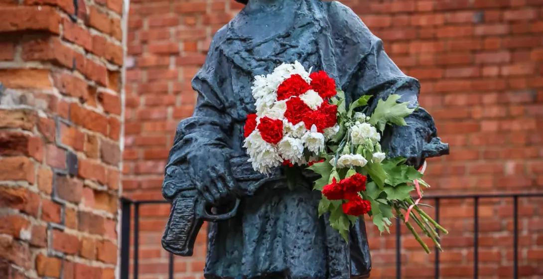 Bronze statue of a child soldier in a helmet holding red and white flowers, against a brick wall.