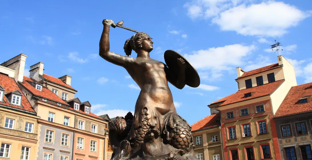 Bronze statue of a mermaid holding a sword with a small bird perched on it, and a shield, against historic buildings and b...