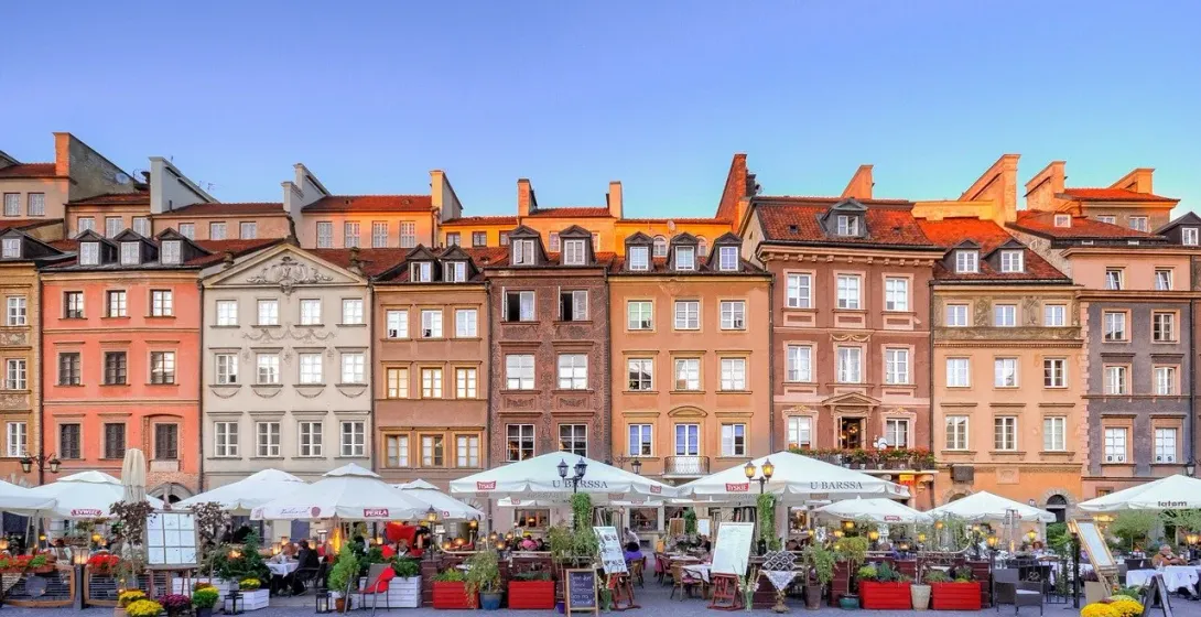Colorful historic buildings with red roofs overlook an outdoor cafe area with white umbrellas and people dining under a bl...