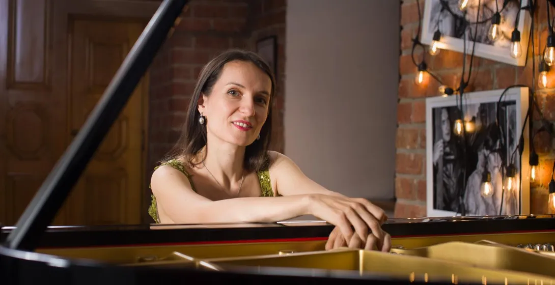 Woman in green top smiling, resting hands on a black grand piano. Brick wall and string lights in the background.