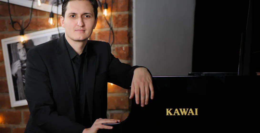 Man in a black suit leaning on a black Kawai piano in a room with a brick wall and string lights in Warsaw.