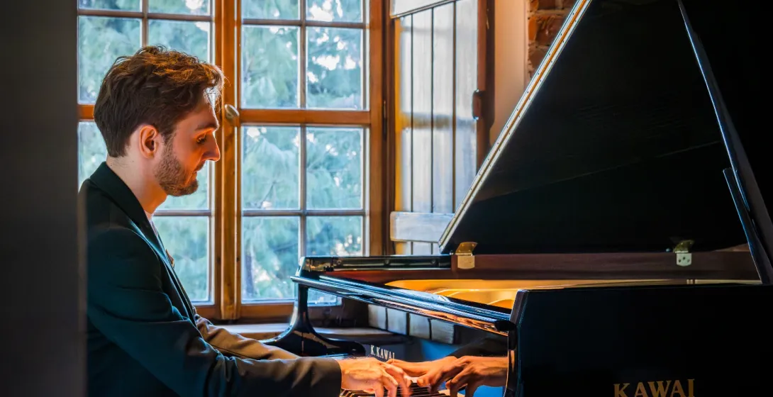 Man in a suit playing a black Kawai grand piano next to a window with wooden shutters.