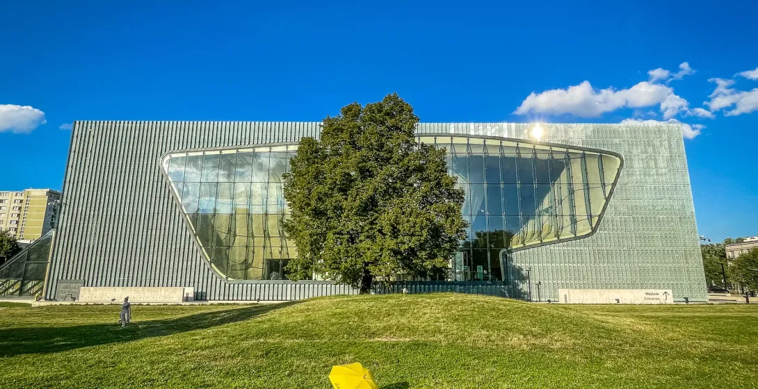 Modern museum building with a wavy glass facade and metal panels, a large green tree, and a yellow umbrella on a grassy mo...