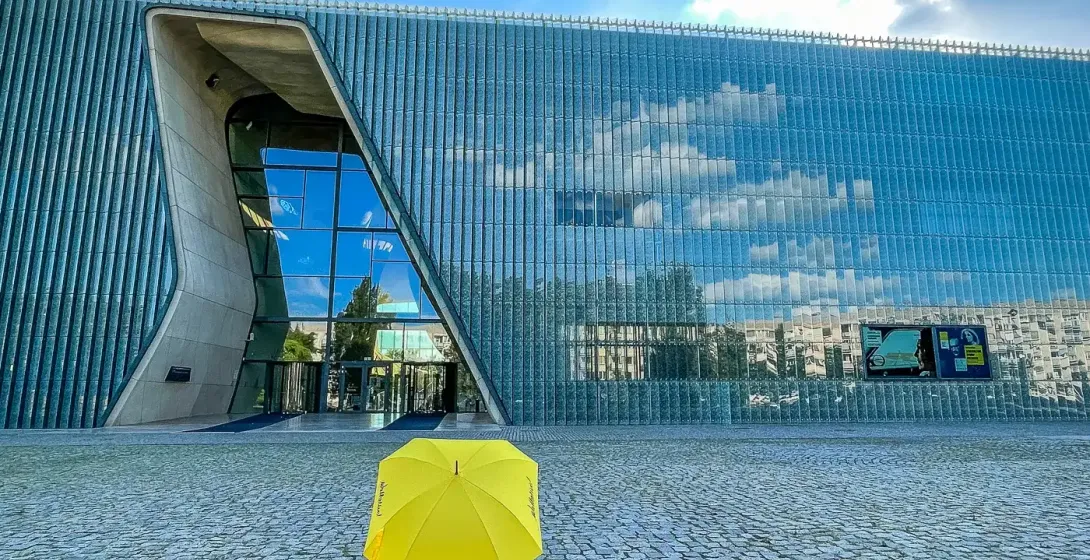 Yellow Walkative umbrella on a cobblestone plaza in front of the modern glass exterior of POLIN Museum in Warsaw under a b...