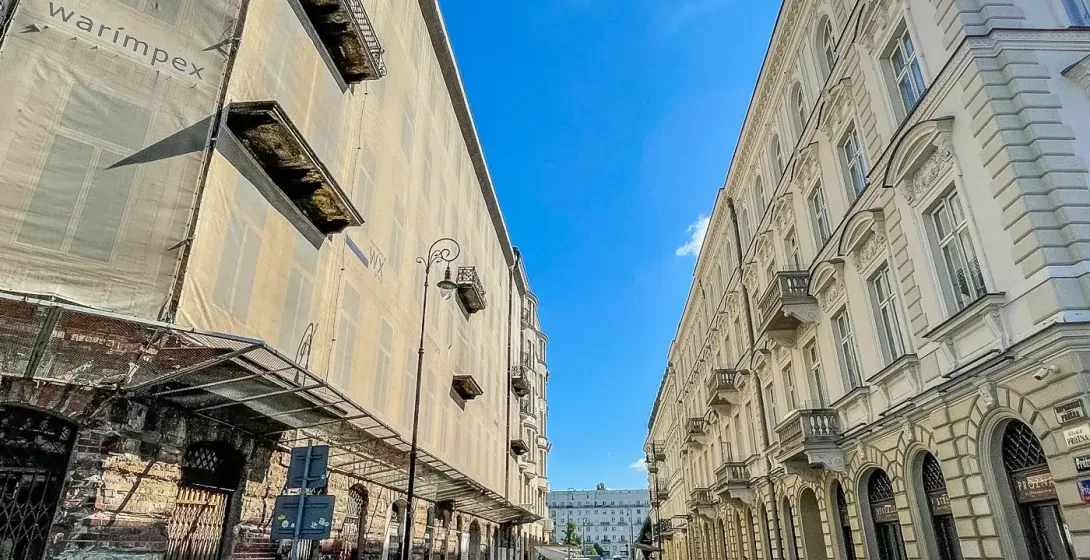Cobblestone street between a scaffolded building with 'warimpex' and an ornate historical building with balconies under bl...