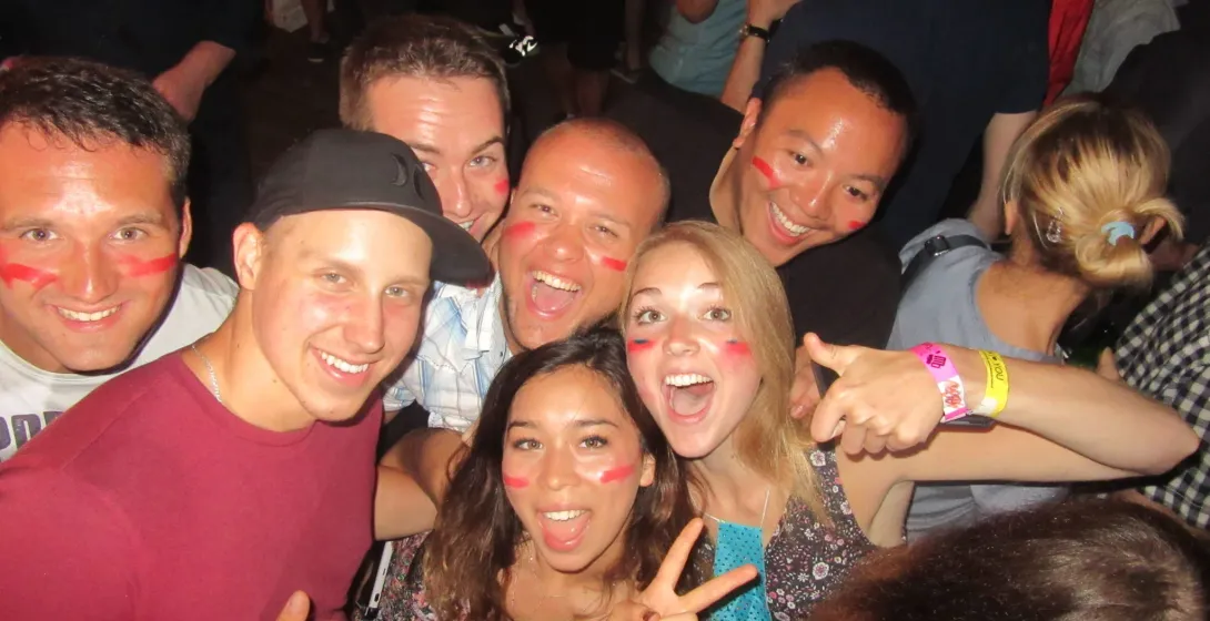 A diverse group of young people smiling, many with red face paint on their cheeks, posing closely together at a lively ind...