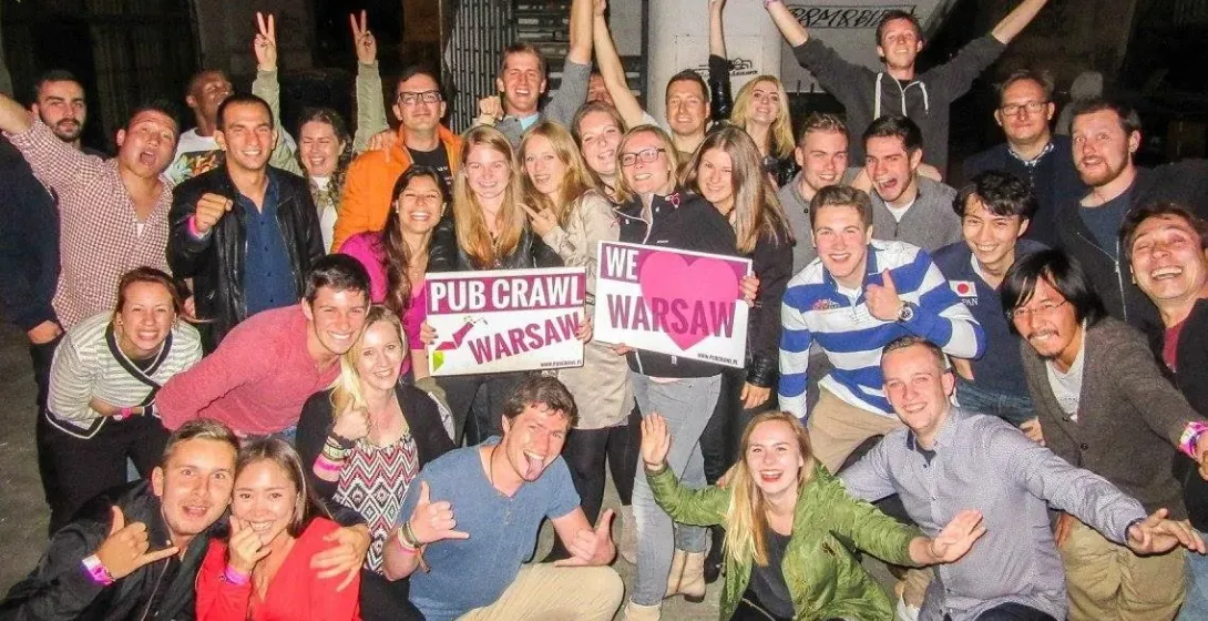 A large group of diverse young adults smiling, posing, and holding "Pub Crawl Warsaw" and "We love Warsaw" signs in an urb...