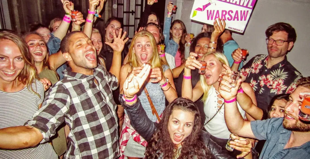 Group of young adults smiling, laughing, and holding shot glasses, one holding a 'PUB CRAWL WARSAW' sign.