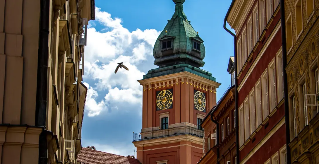 View up a narrow street towards the Royal Castle clock tower in Warsaw with a green dome and golden clock faces, a bird fl...