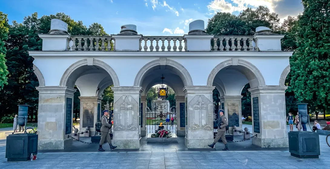 Two uniformed guards march in front of the white stone arcades of the Tomb of the Unknown Soldier in Warsaw under a blue sky.