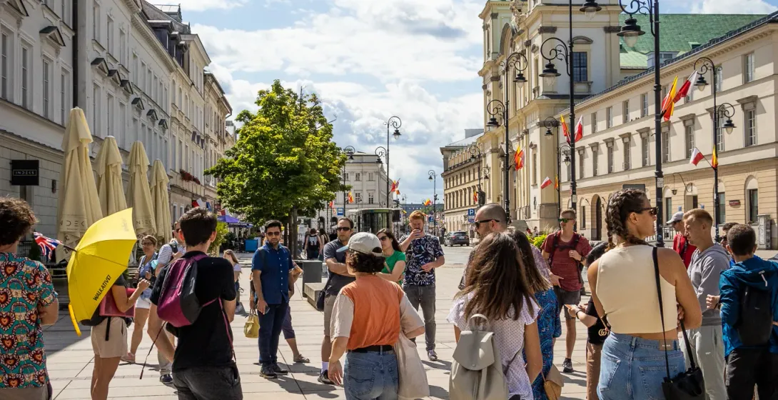 A group of people gathered on a paved street in Warsaw with a historic church and other buildings in the background under ...