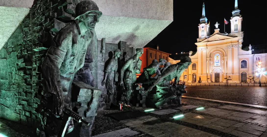Bronze monument depicting figures of Warsaw Uprising soldiers under green light at night, with a brightly lit church behind.