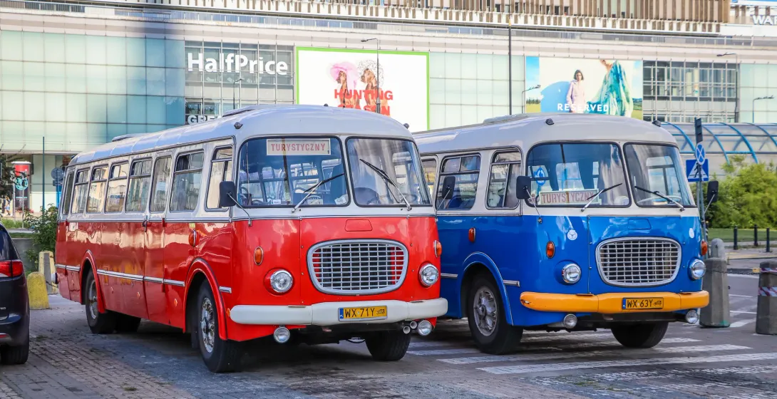 Two vintage Polish tourist buses, one red and one blue, parked on a city street in front of a modern building.