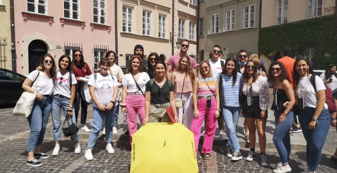 A group of young adults poses on a cobbled street with a yellow Walkative umbrella. Colorful historic buildings in backgro...