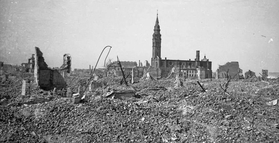 Black and white photo of a city devastated by war, showing a vast field of rubble in the foreground and a damaged church w...