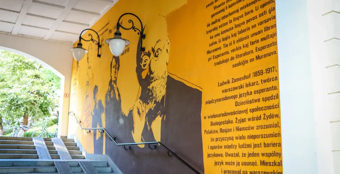 Large yellow mural depicting Ludwik Zamenhof and text, next to a concrete staircase with handrails and lamps.