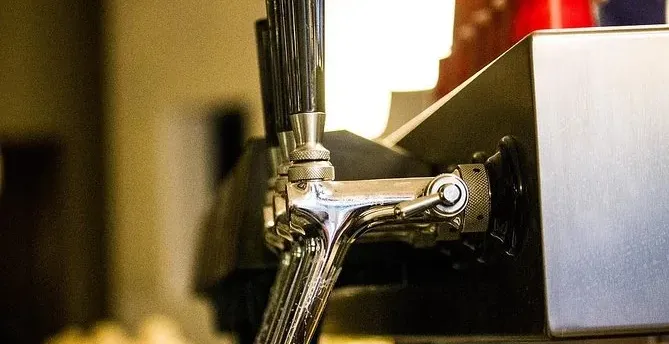 Close-up of three chrome beer taps with black handles on a bar. The background is blurred, showing warm lighting.