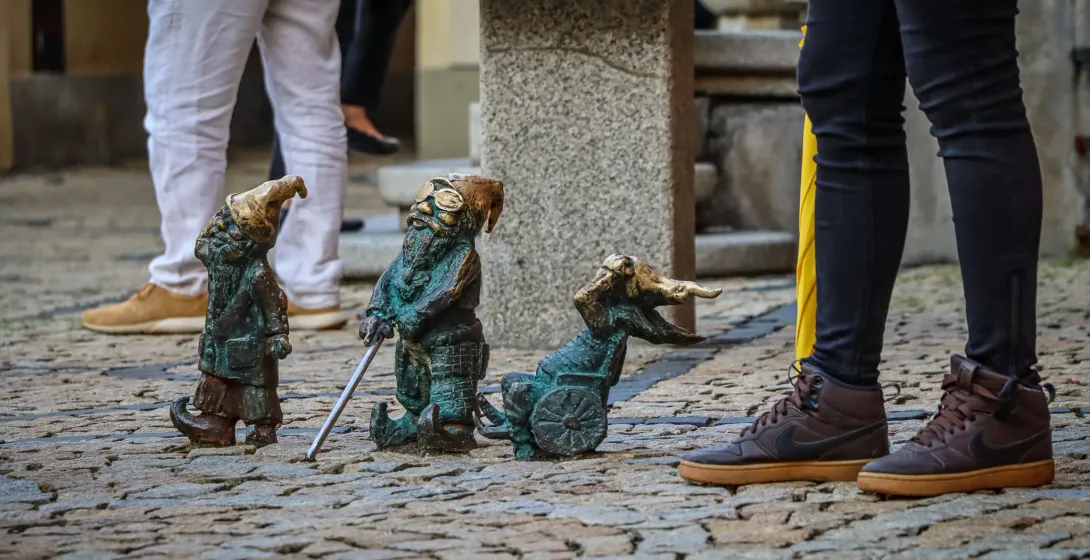 Three bronze dwarf statues on a cobblestone street, with parts of people's legs visible around them. One dwarf holds a staff.