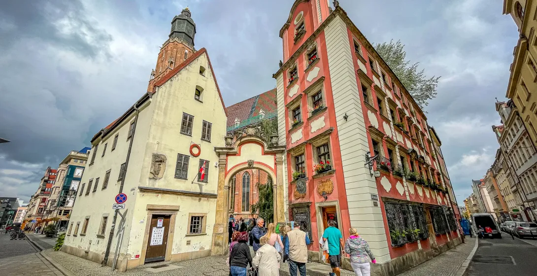 People walk on a cobblestone street in Wroclaw's Old Town, past historic yellow and red buildings, and an archway leading ...