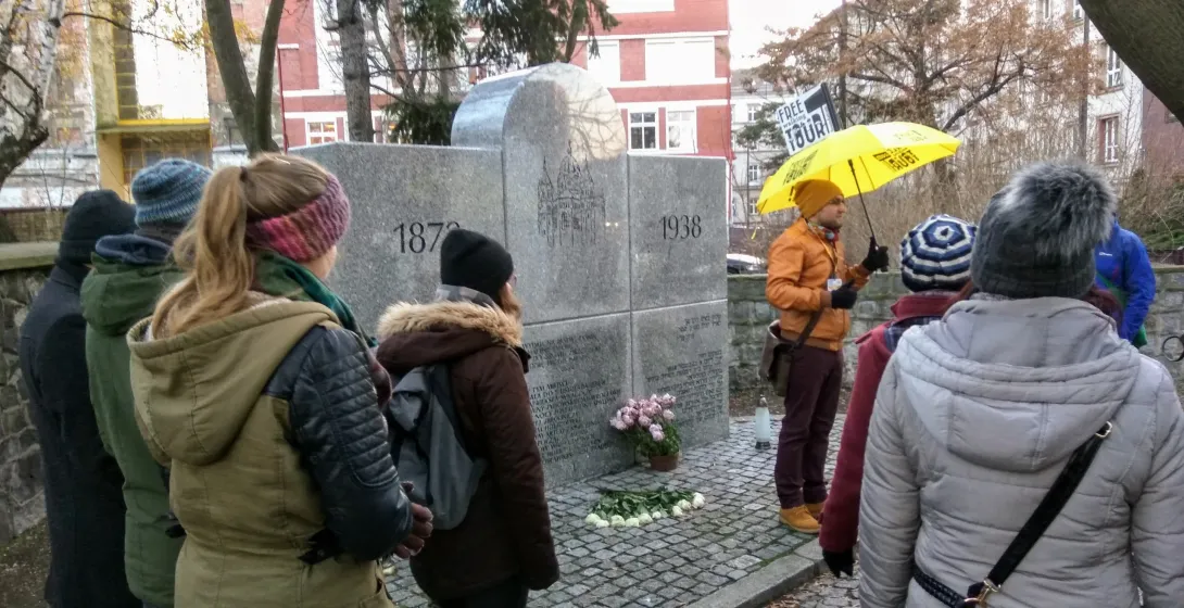 Stone monument commemorating the New Synagogue with dates 1870 and 1938, attended by a tour group with guide.