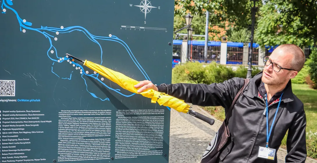 Man pointing with a yellow umbrella at an information board displaying a map of Wrocław's Floodway System on a sunny day.