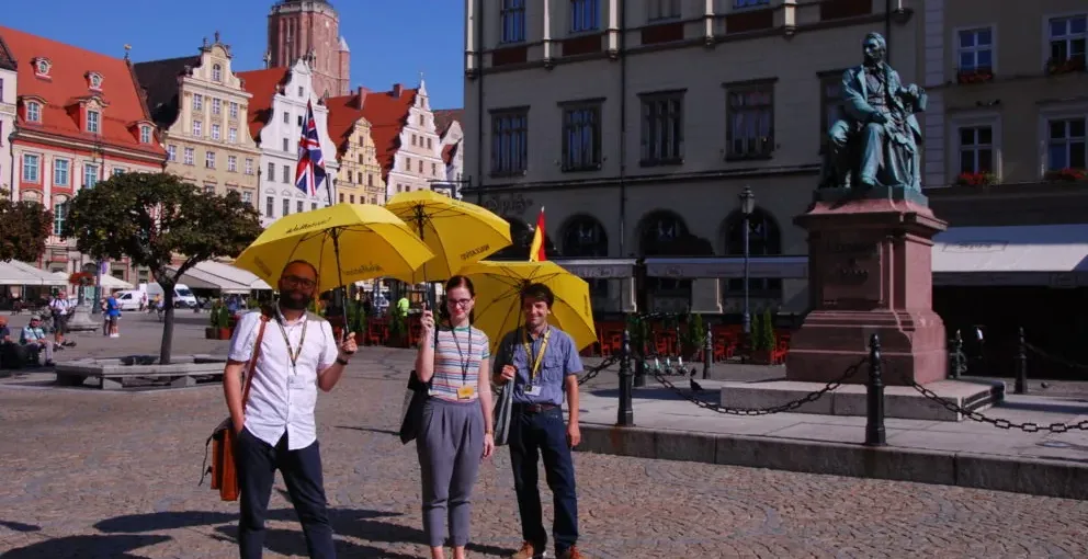 Three people with yellow umbrellas stand on a cobblestone square in front of historic buildings and a statue on a sunny day.