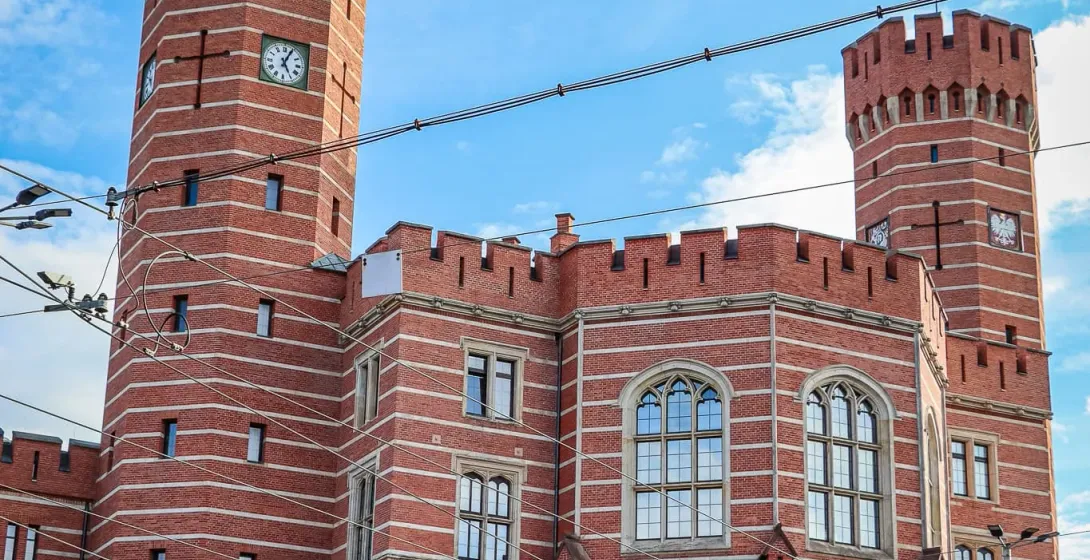 Historic red-brick building with two towers, each featuring a clock face, under a blue sky with white clouds in Wroclaw.