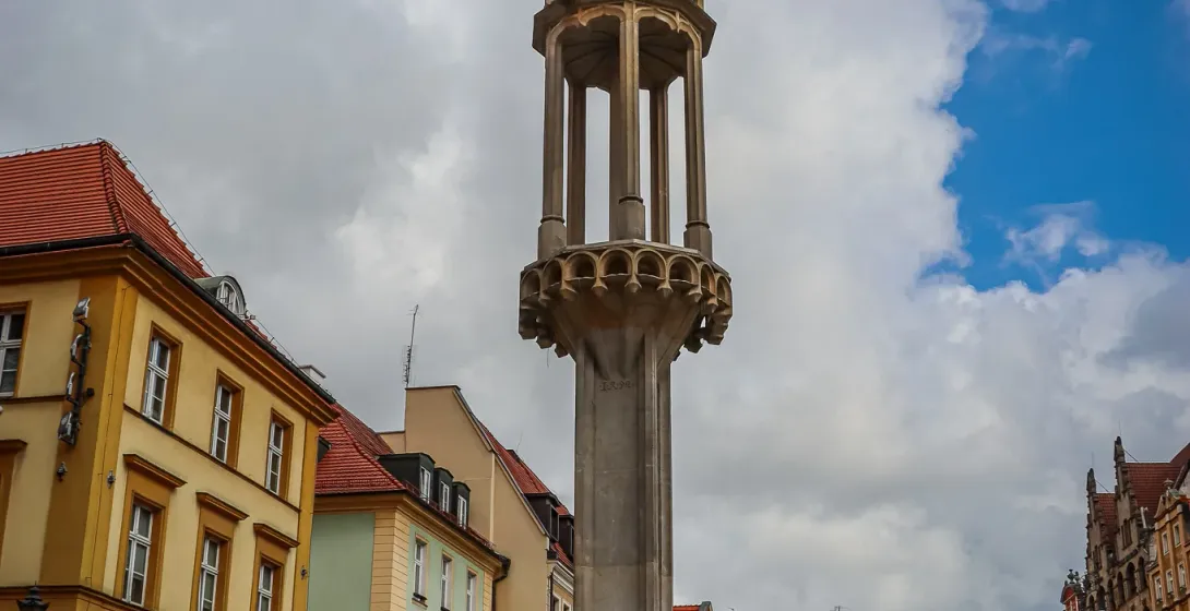 Tall stone pillory with an ornate spire and a figure on top, surrounded by colorful historic buildings in a European city ...