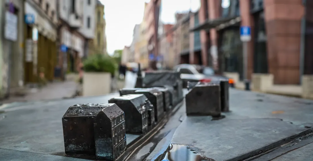 Close-up of a dark metal miniature model of city buildings with water puddles, blurring into a real street scene in the ba...