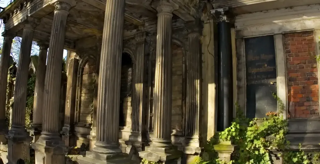 Stone mausoleum with classical columns, intricate stonework, and green ivy, surrounded by fallen yellow autumn leaves unde...