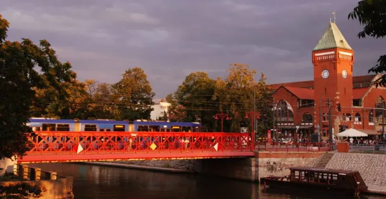 Red metal bridge over a river with a blue train crossing, next to a red brick market hall with a clock tower at sunset.