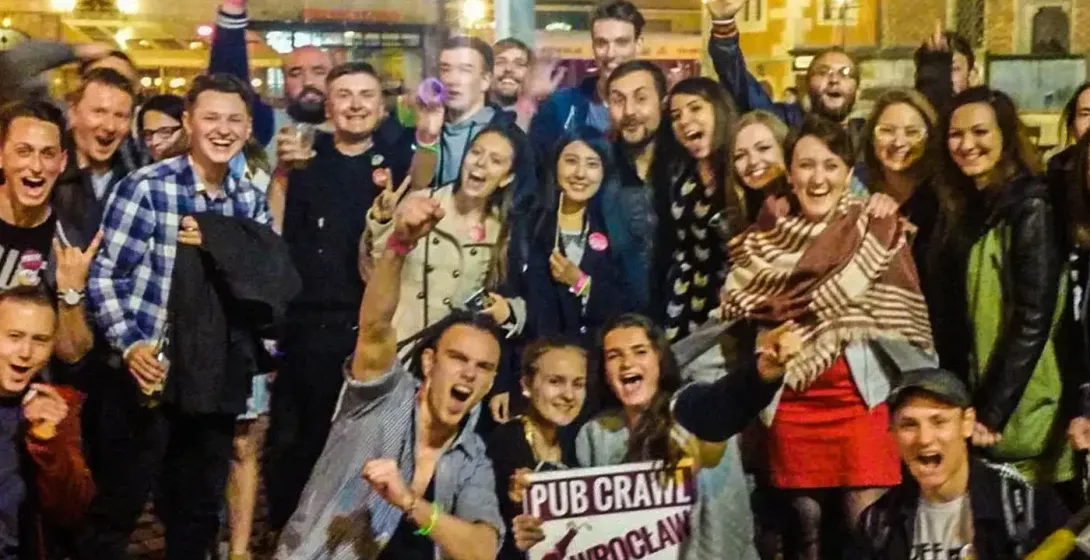 Group of cheerful people at a night pub crawl in Wrocław, with historic buildings in the background. A sign reads "Pub Cra...