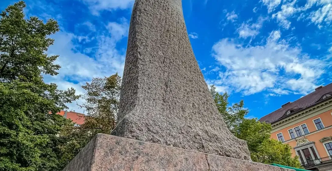 Tall, twisting abstract stone monument with a rough texture, on a granite base, against a bright blue sky with white clouds.