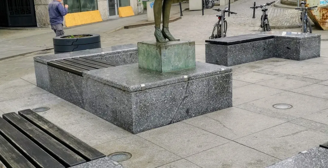 Bronze statue of a woman with a globe as a skirt, granite benches, and a metal plaque with religious symbols on a city str...