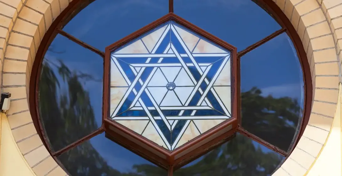 Round stained glass window with a blue and white Star of David design, set in a yellow brick arch above a brown wooden frame.