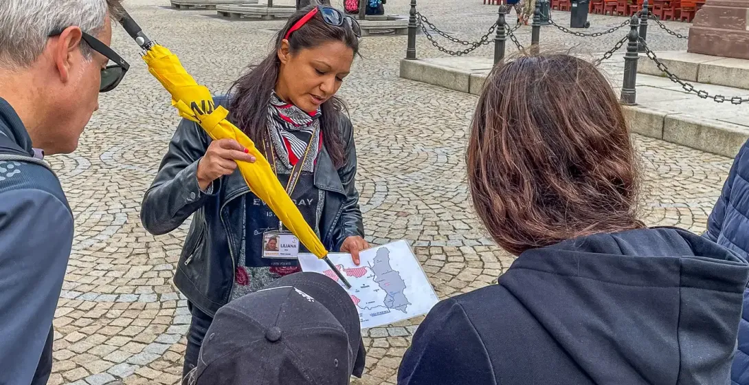 Female tour guide with yellow umbrella and map explains to three tourists on a cobblestone square. Colorful historic build...
